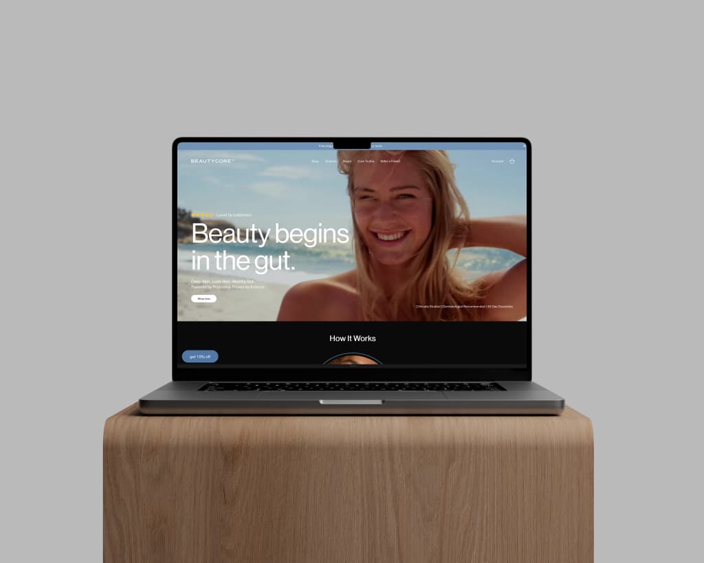 Laptop on a wood pedestal showing the BeautyCore homepage: beach hero, “Beauty begins in the gut.” headline, Shop now button, and How It Works section